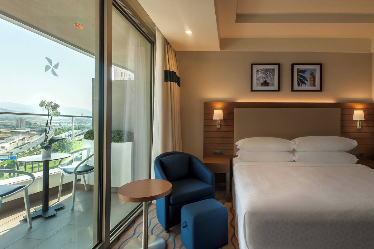 Habitación de hotel con cama, una silla, una mesita y balcón con vistas a la ciudad.