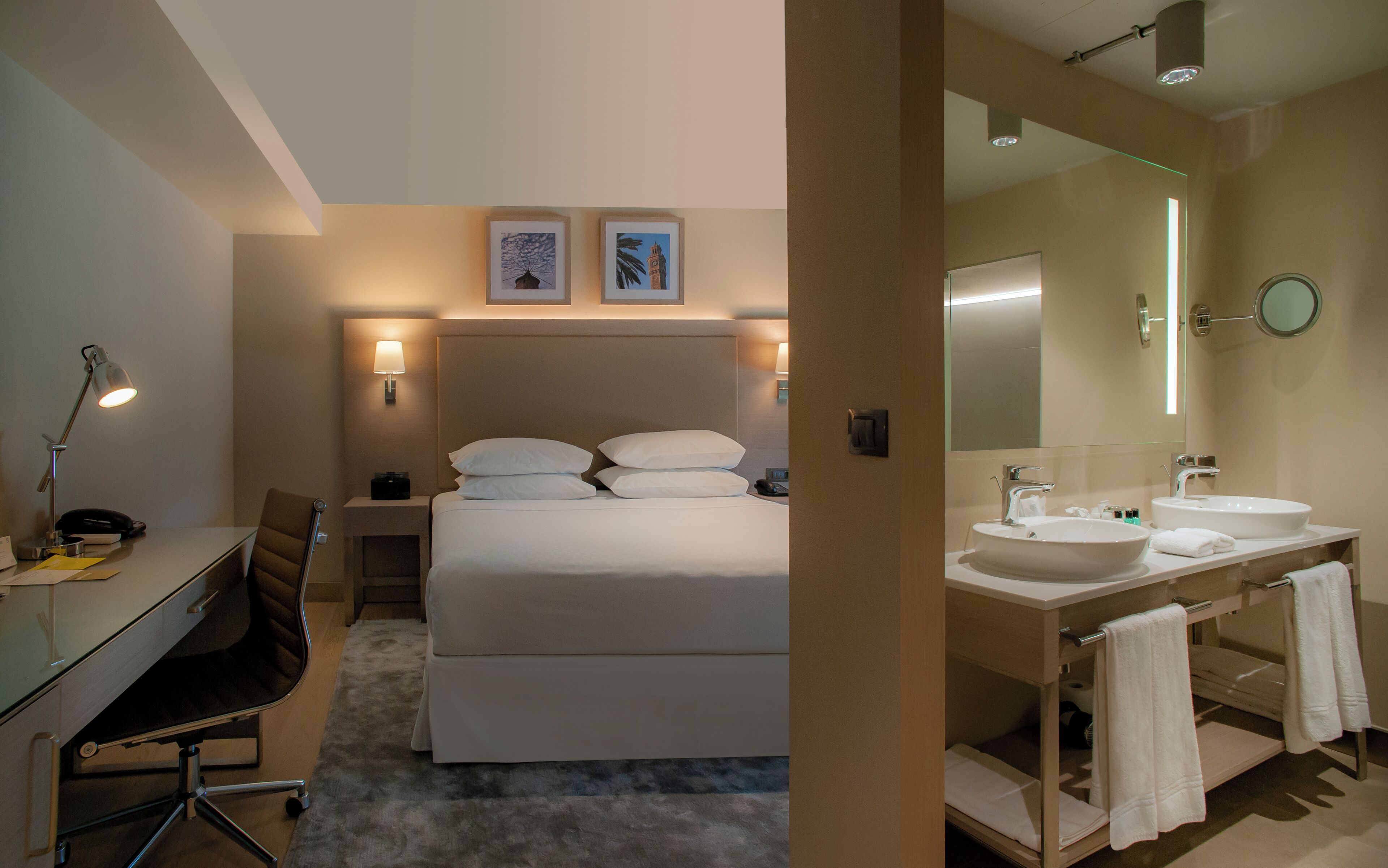 Quarto de hotel com cama, abajures, escrivaninha com cadeira, luminária de mesa, telefone, espelho e banheiro com pia e porta-toalhas.