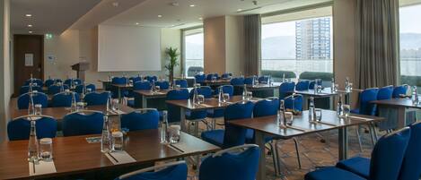 Un salón de conferencias con varias mesas y sillas azules, equipado con botellas de agua y vasos, y una pantalla grande al frente.