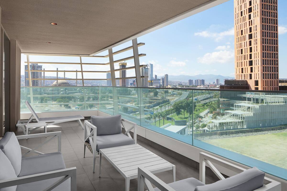 Un balcón moderno con mobiliario blanco y barandilla de cristal, con vistas a la ciudad.
