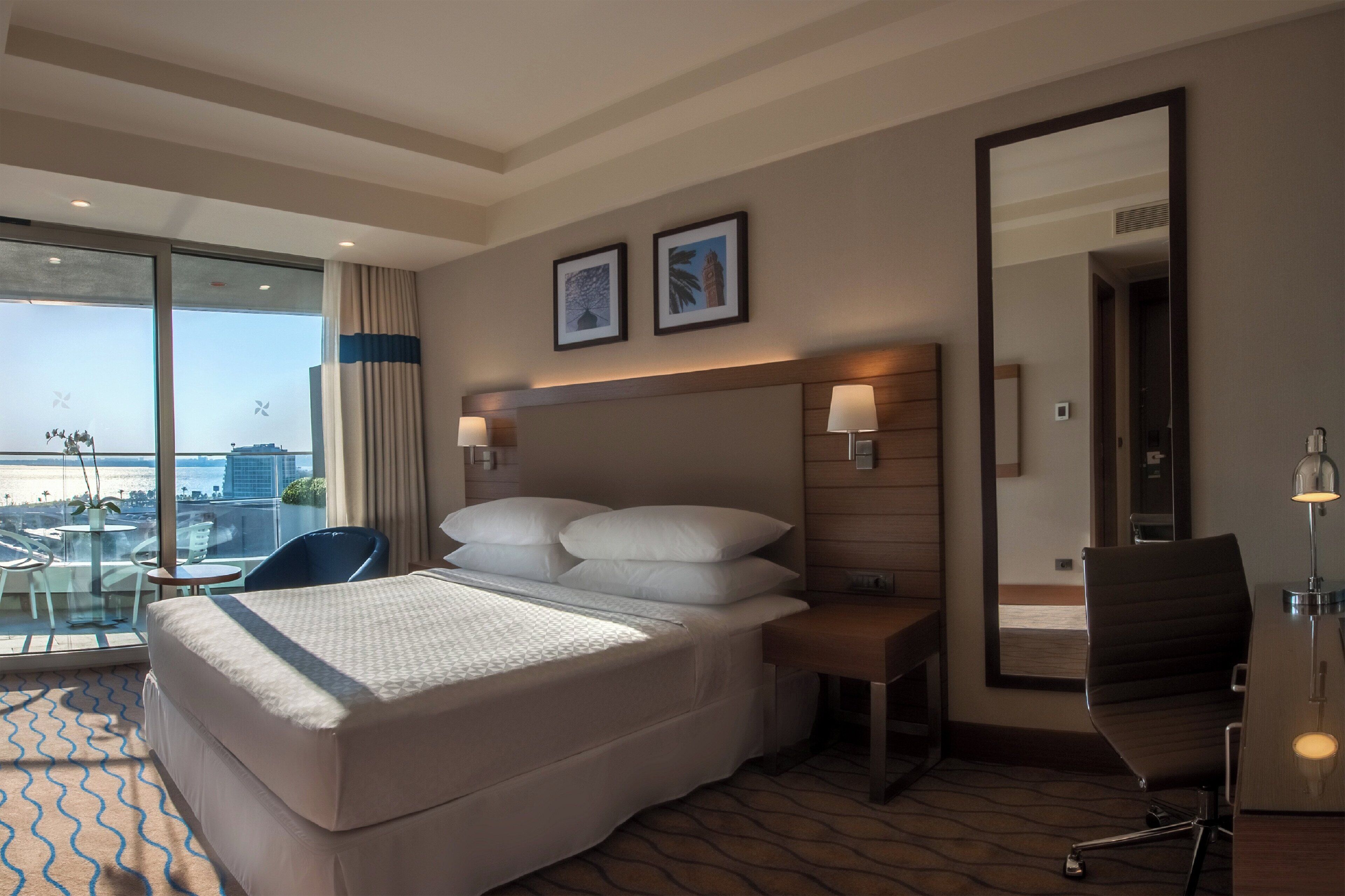 Une chambre d’hôtel avec un grand lit, un bureau, une chaise, une lampe de bureau, un miroir et une vue sur la ville grâce à de grandes fenêtres.