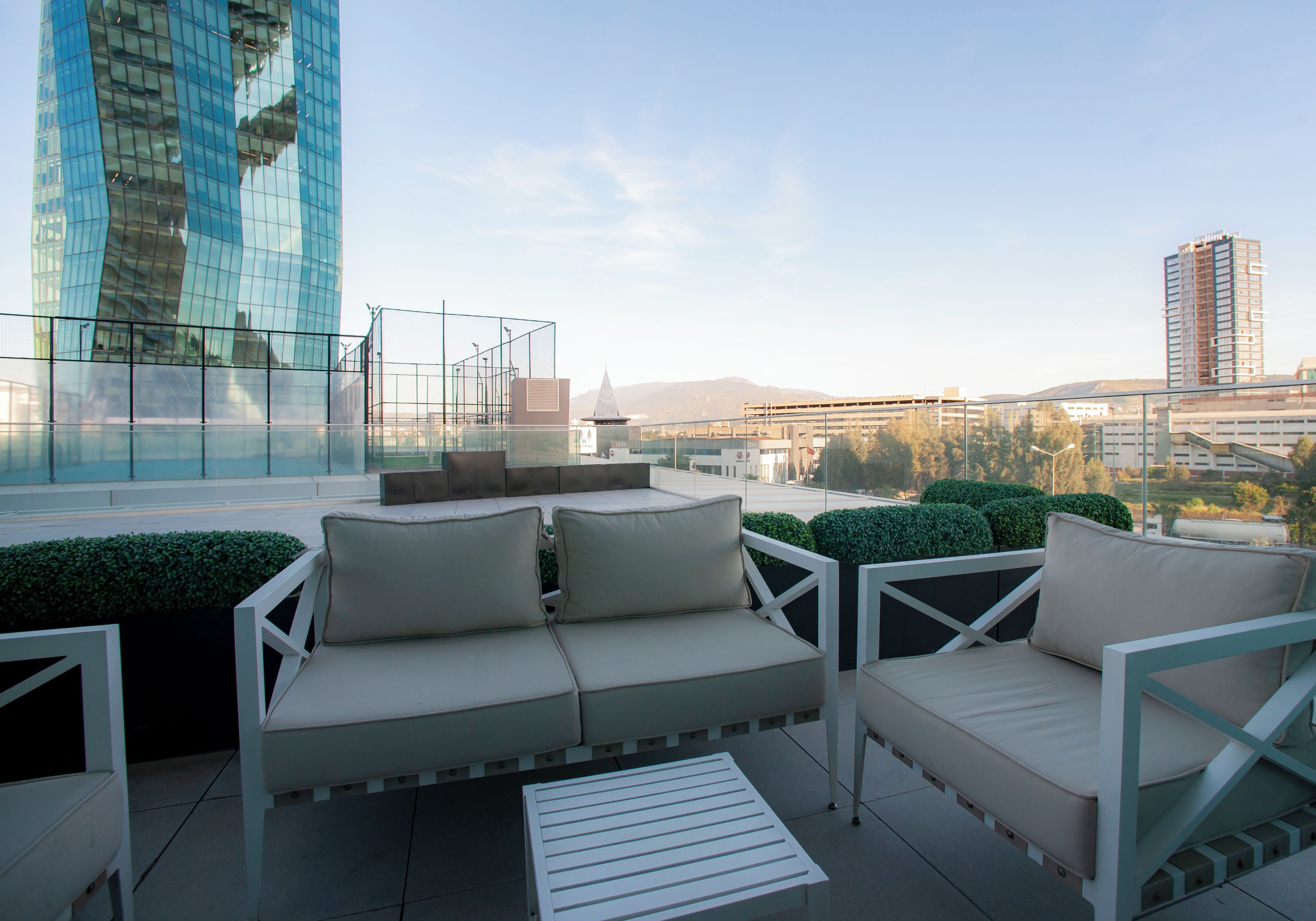 A rooftop terrace with a white outdoor seating area, a glass building in the background, and a view of the cityscape.