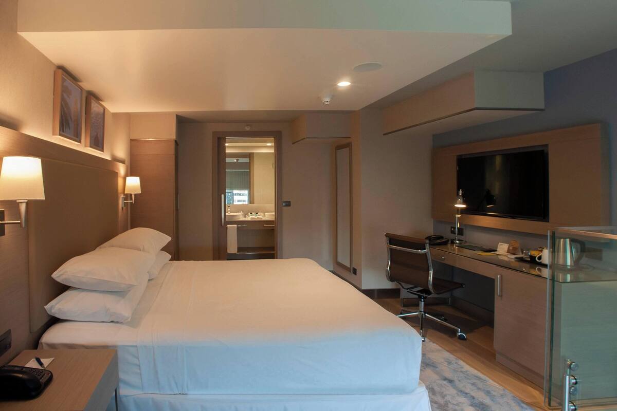 Habitación de hotel con cama, escritorio, televisor y baño visible a través de una puerta abierta.