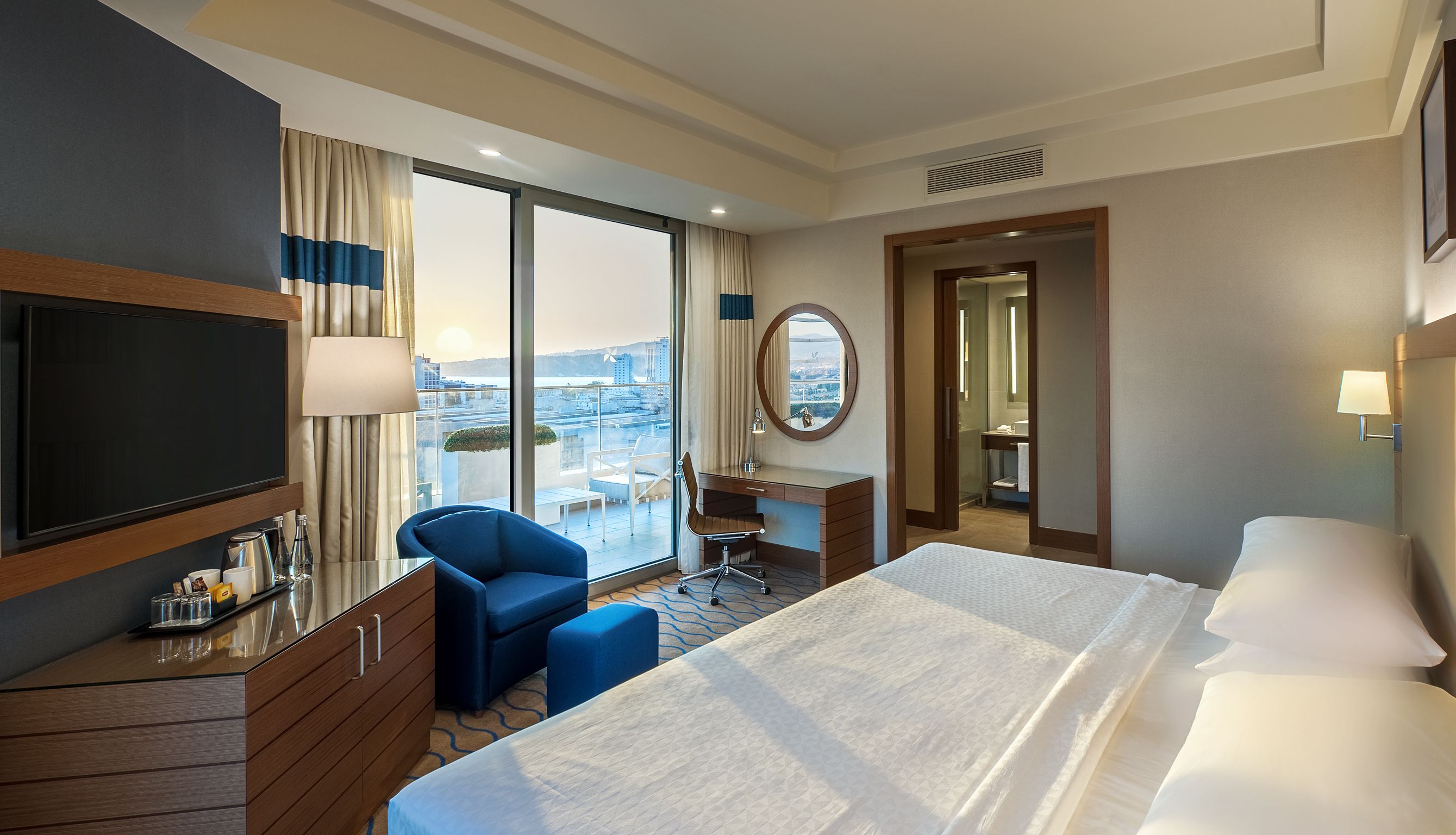 A hotel room with a large bed, a desk with a chair, a television, and a view of the city through large windows.