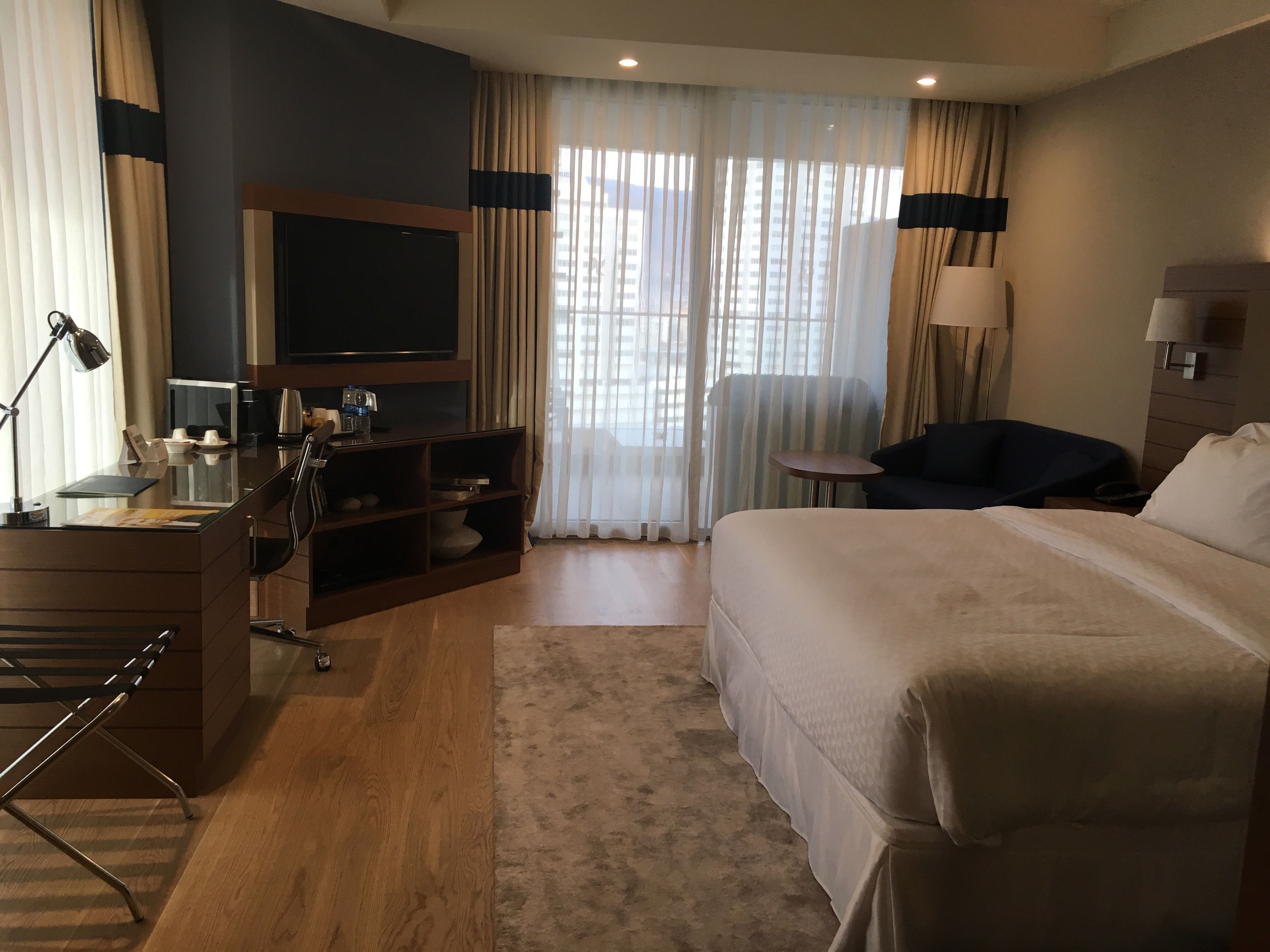 A hotel room with a large bed, a desk with a computer, a TV, and a seating area with a chair and a small table.