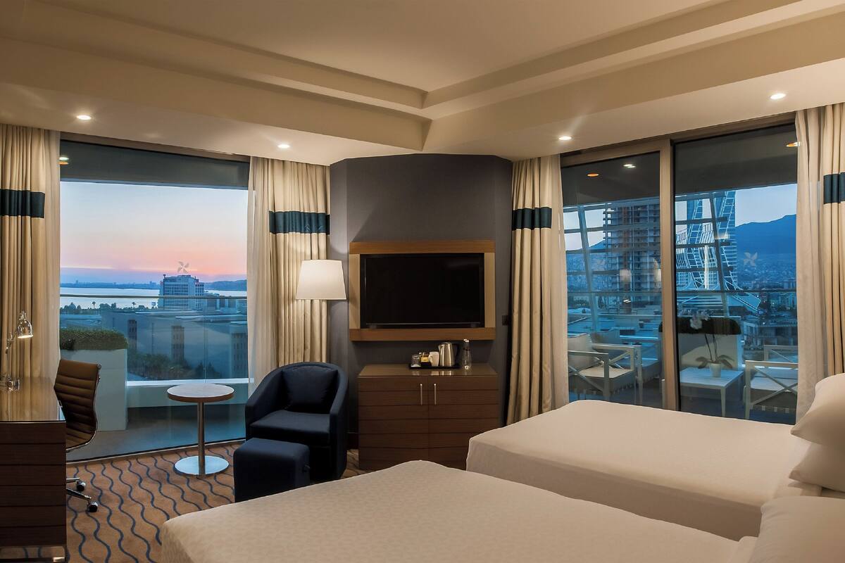 Habitación de hotel con dos camas, televisión, escritorio y una silla. Se ve la ciudad a través de amplias ventanas.