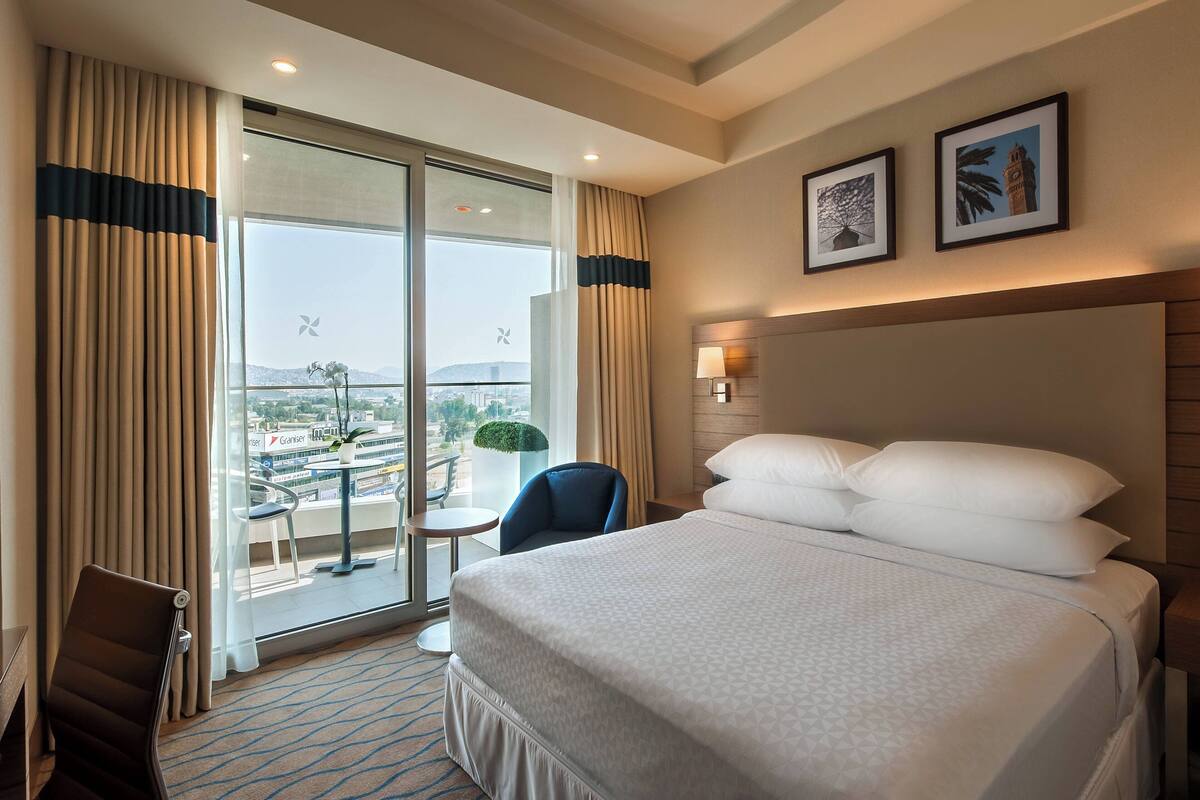 Habitación de hotel con una cama grande, una silla, una mesita y vistas a la ciudad a través de un ventanal amplio.