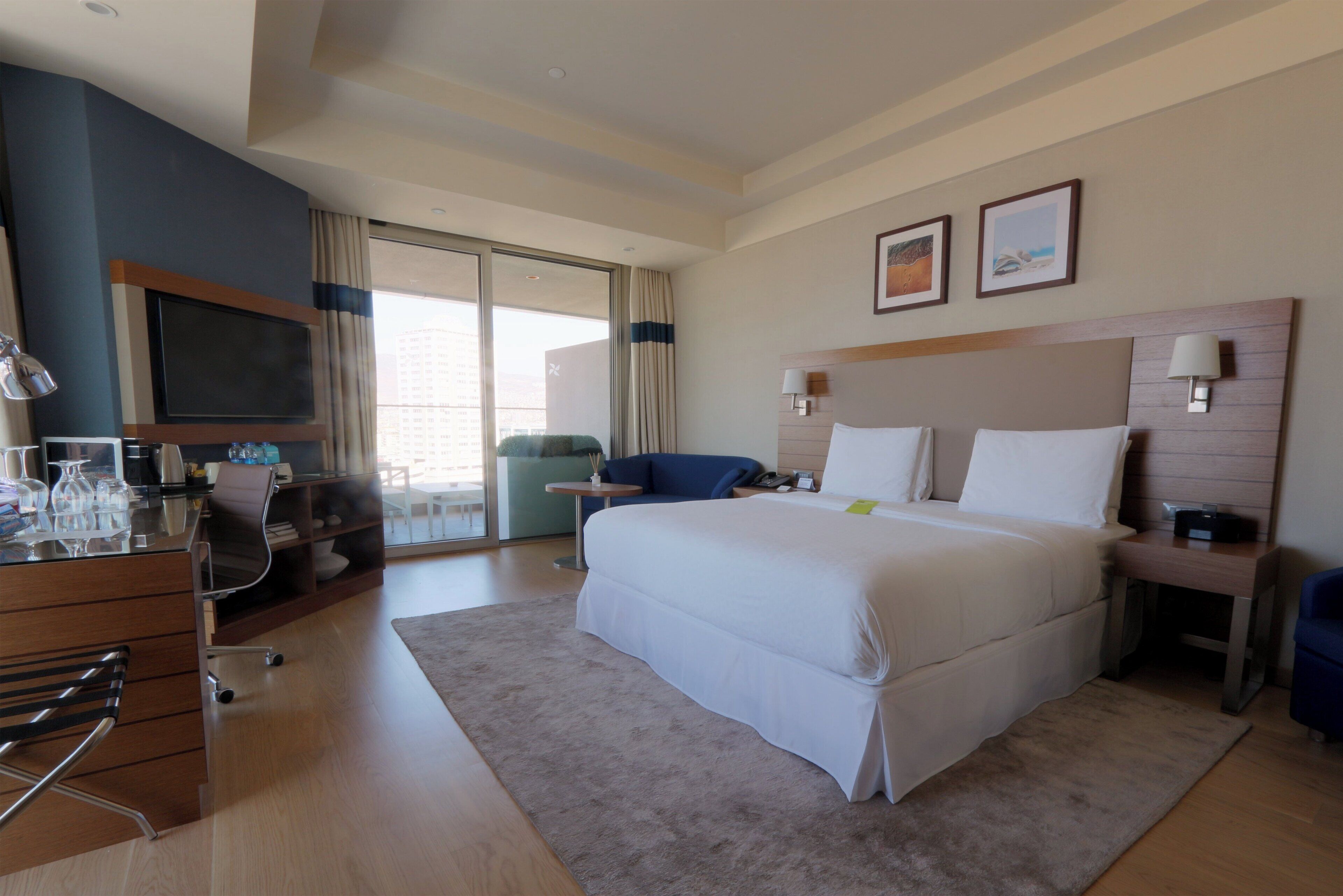 A hotel room with a large bed, a desk with a computer, a TV, and a view of the city.