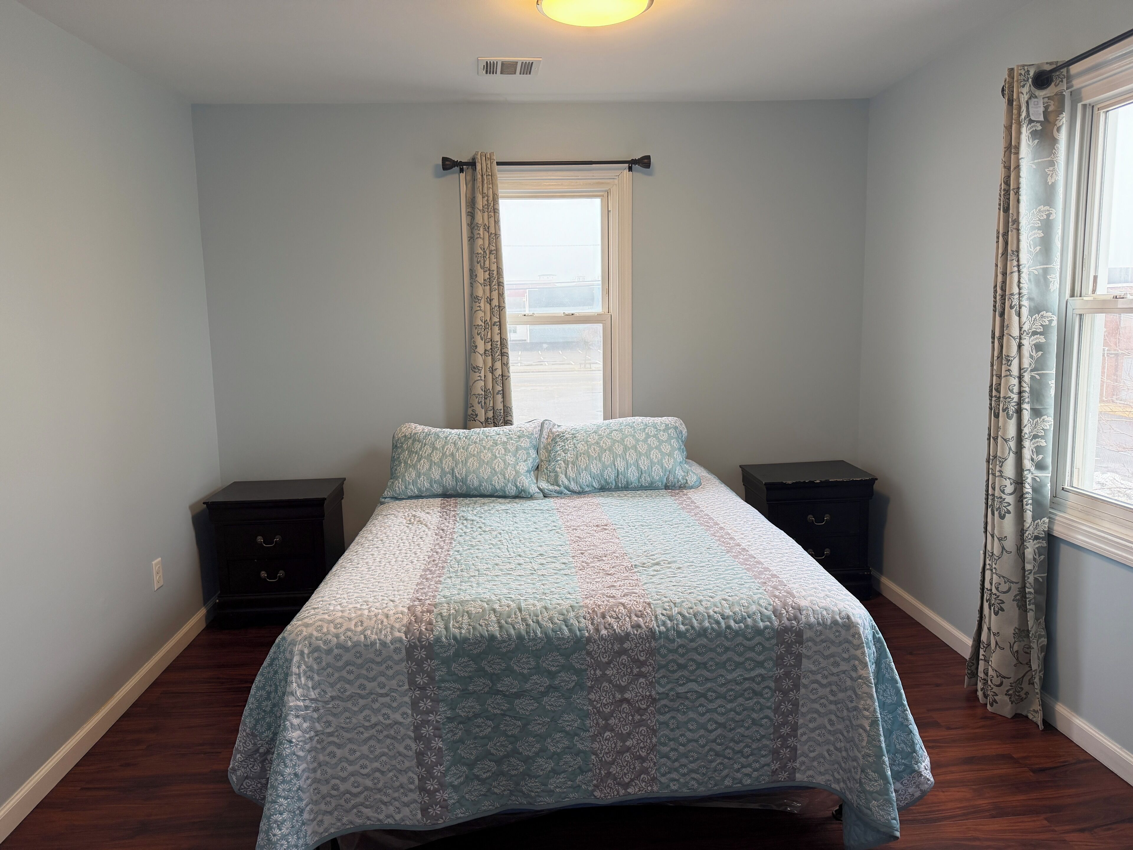 A bedroom with a bed, two nightstands, a window with curtains, and a ceiling light.