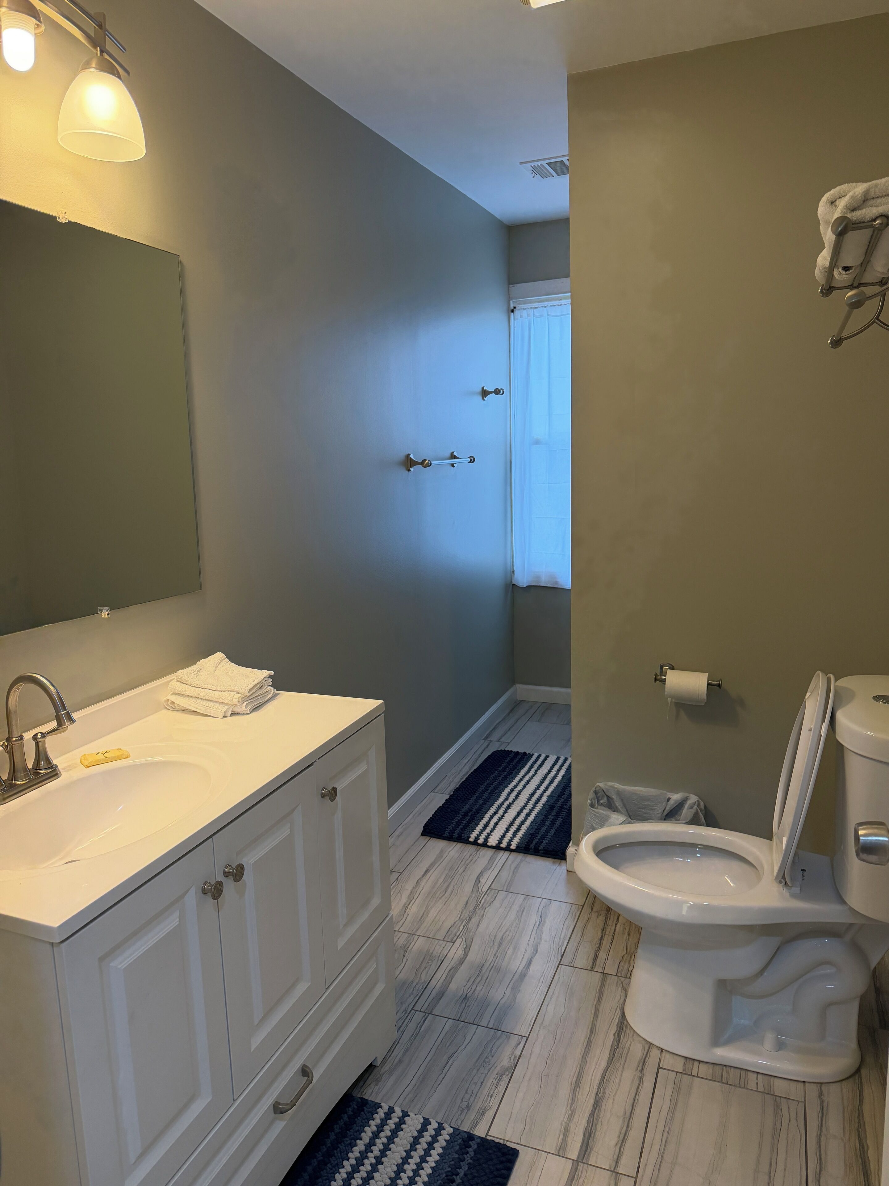 A bathroom with a white sink, mirror, and toilet. There is a towel rack above the sink and a window with a curtain.