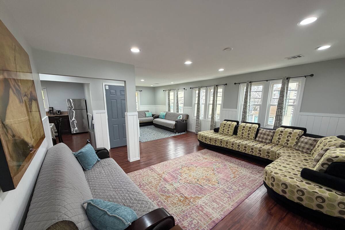 A spacious living room with a sofa set, a coffee table, and a rug. There is a kitchen area in the background with a refrigerator and a stove.