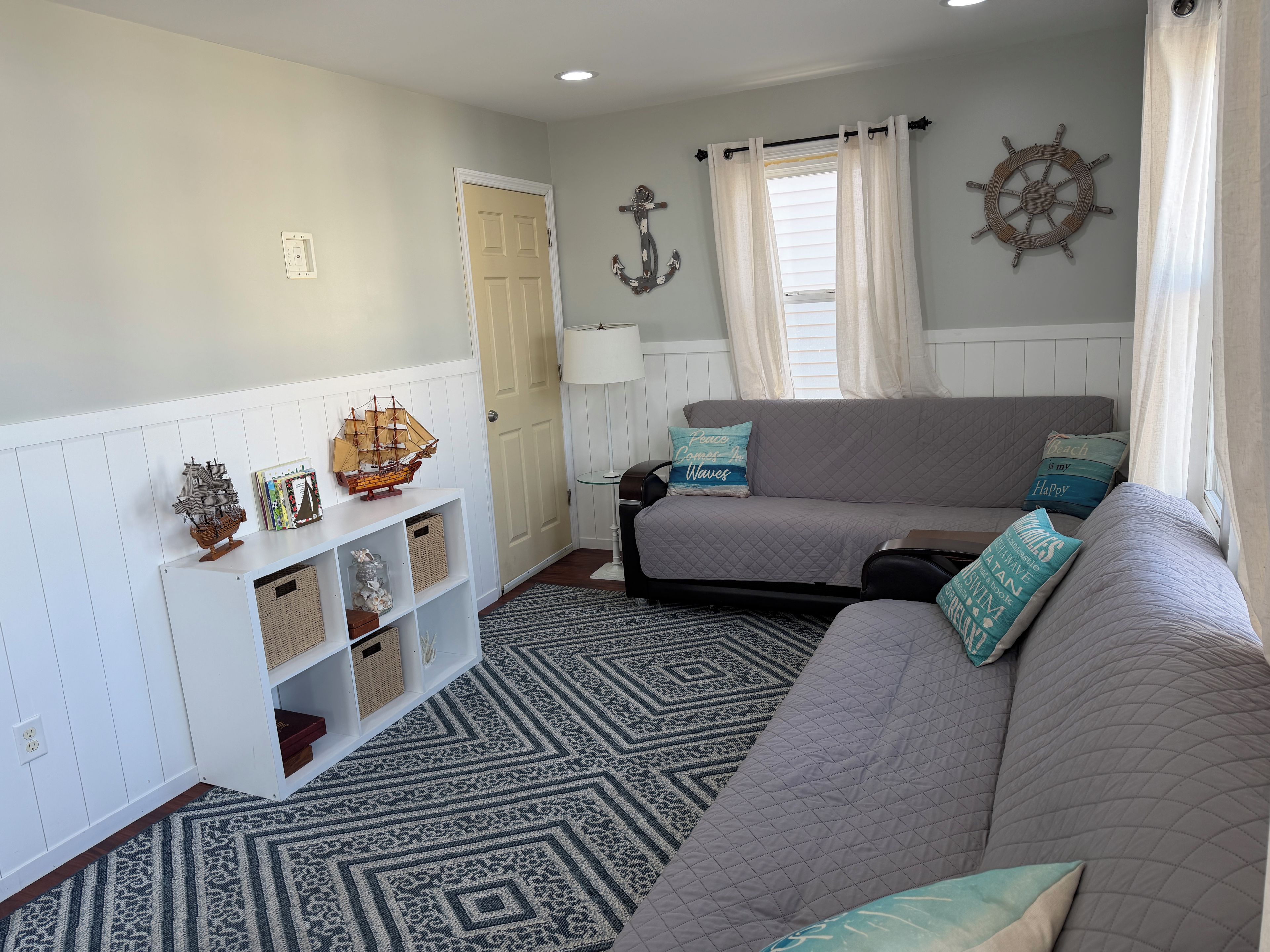 Ein gemütliches Wohnzimmer mit grauem Sofa, einem gemusterten Teppich und maritimer Dekoration, darunter ein Schiffmodell und ein Steuerrad.