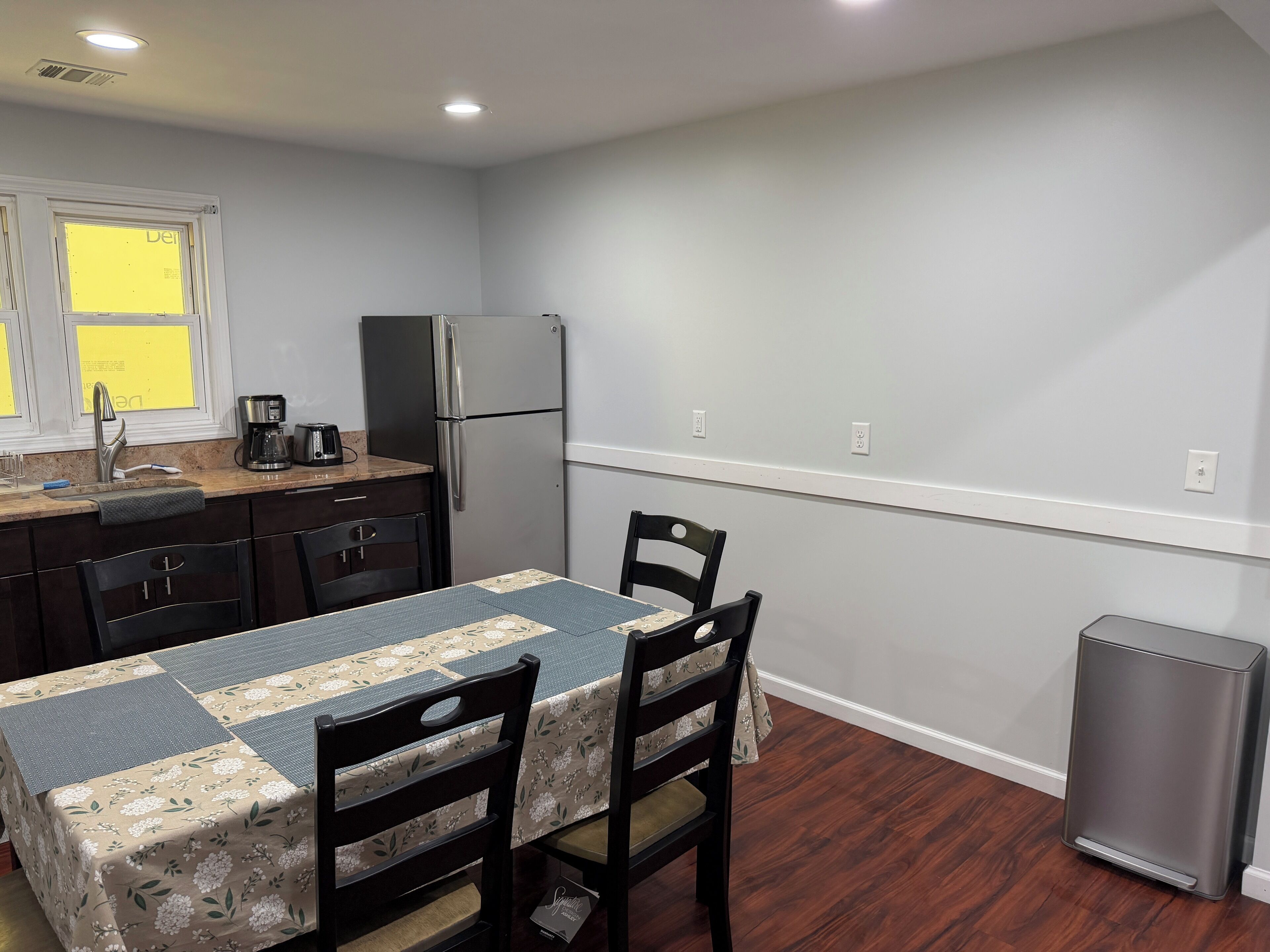 A compact kitchen with a refrigerator, a coffee maker, and a sink. There are two chairs and a table with a floral tablecloth.
