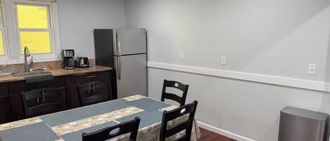 Cocina compacta con refrigerador, cafetera y lavamanos. Hay dos sillas y una mesa con mantel floreado.