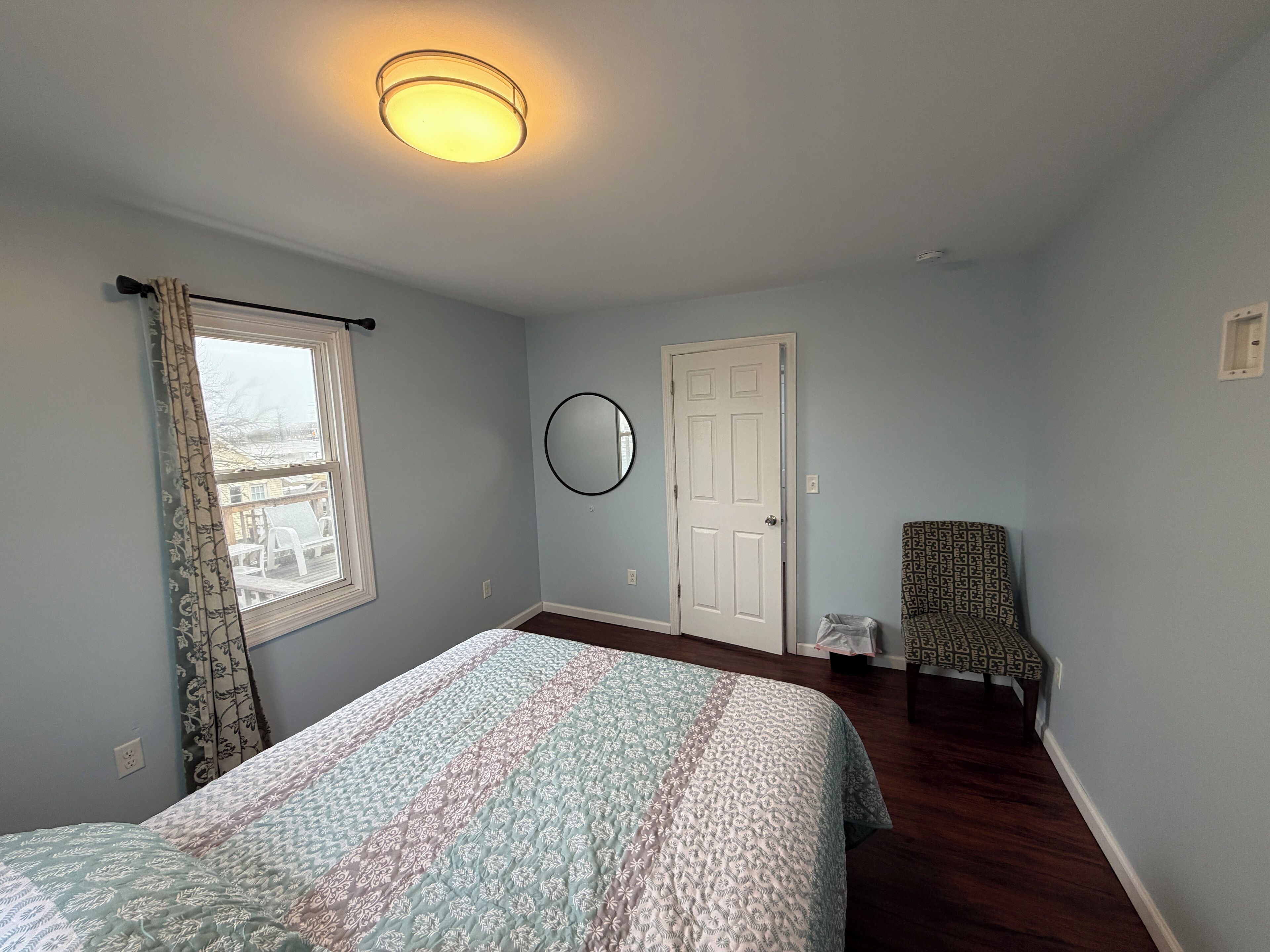 A bedroom with a bed, a window with curtains, a door, a chair, and a round mirror on the wall.