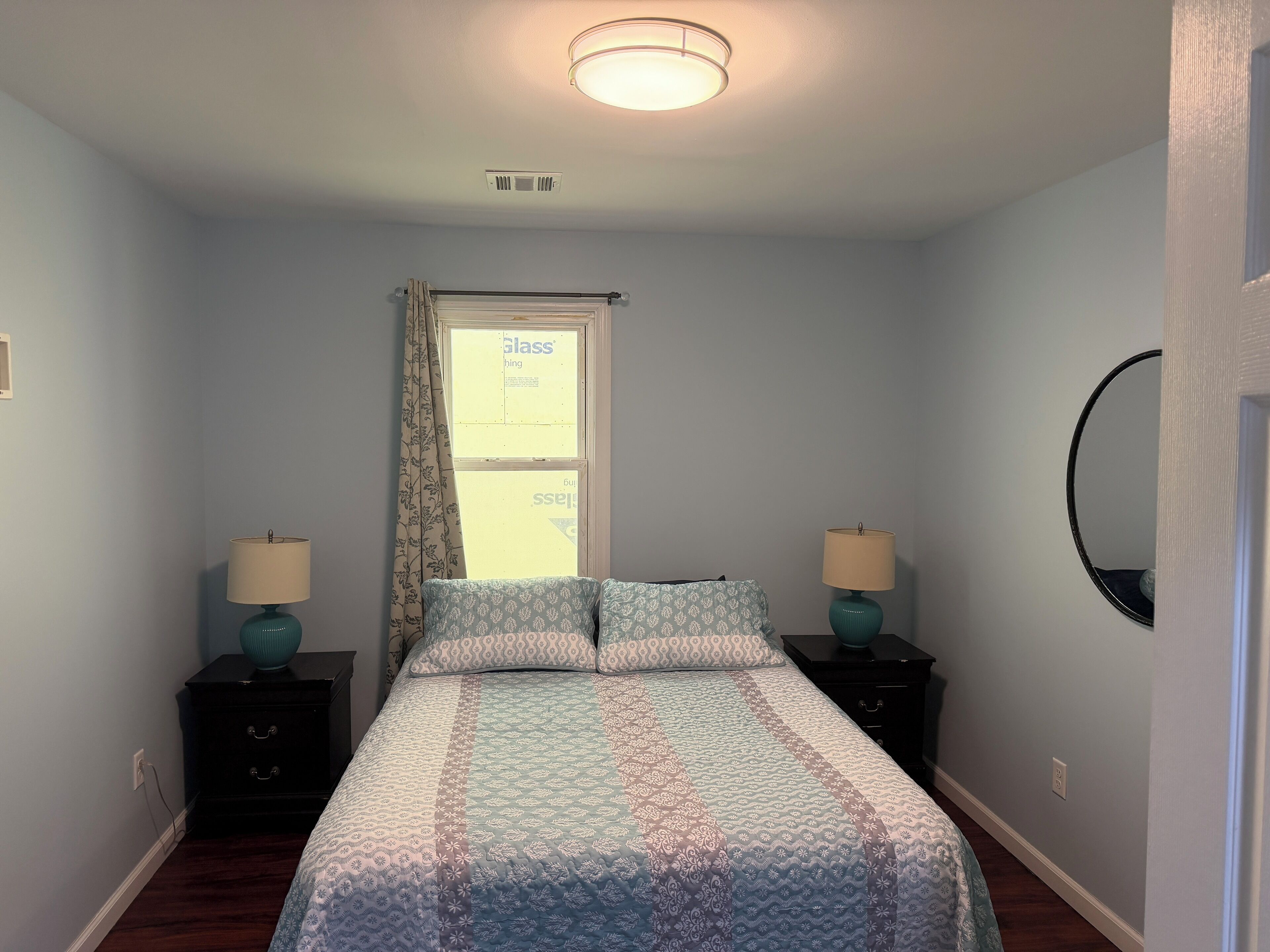 A bedroom with a bed, two bedside tables, a window with curtains, a ceiling light, and a wall-mounted mirror.