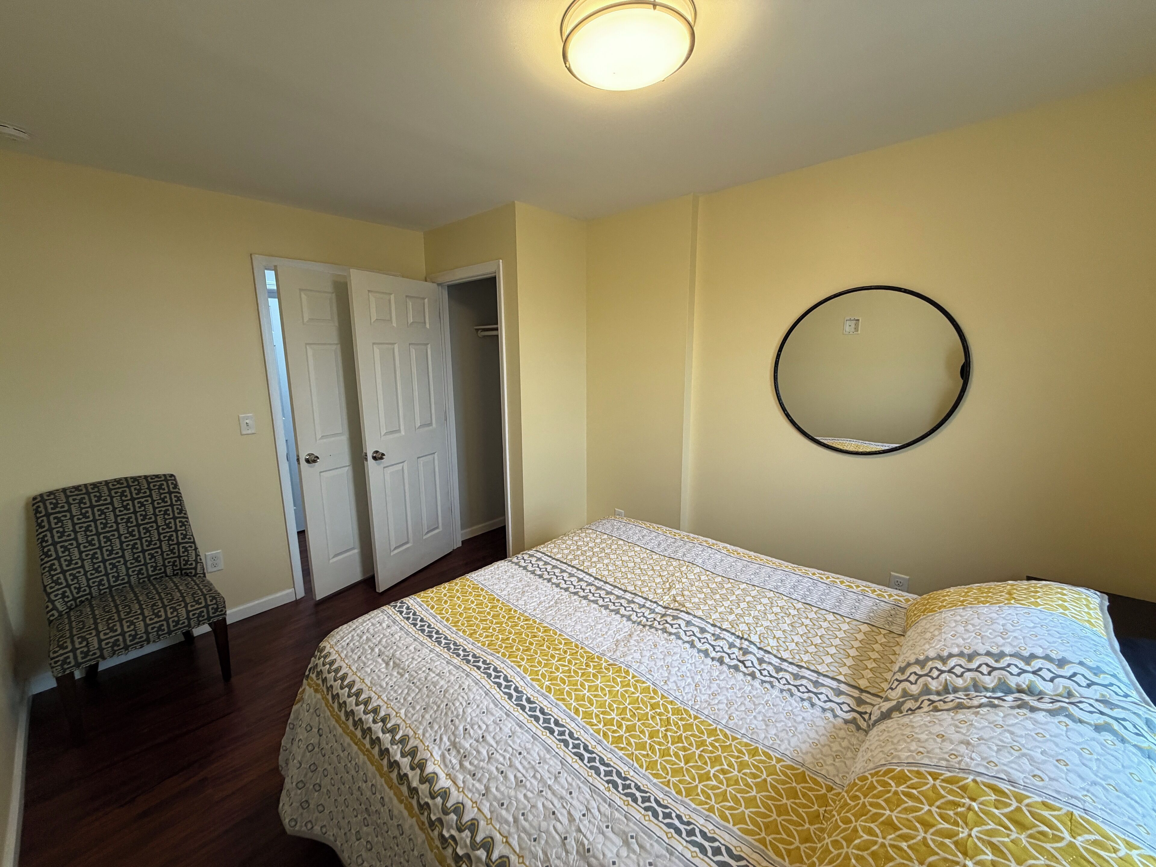 A bedroom with a bed, a chair, a door, and a mirror on the wall.