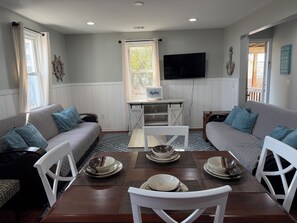Un coin repas avec une table dressée pour quatre personnes, entouré de deux canapés, d’un téléviseur fixé au mur et d’une fenêtre avec des rideaux.