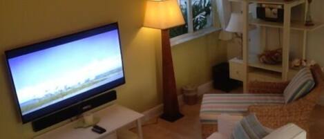 A living room with a flat-screen TV, a ceiling fan, a lamp, a sofa, and a window with a view of greenery.
