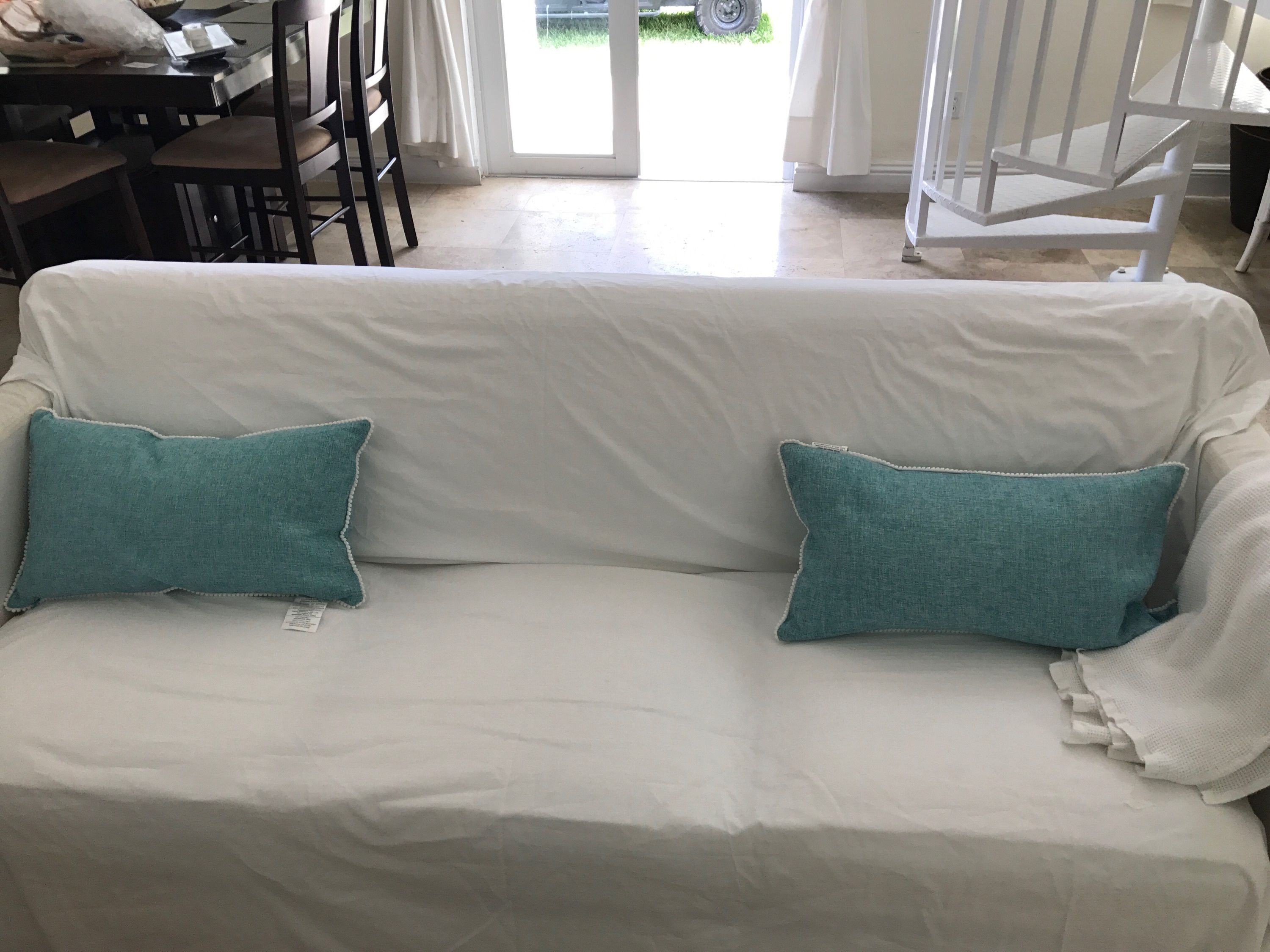 Un sofá blanco con almohadones verde azulado, una mesa de comedor con sillas, y vista al exterior.