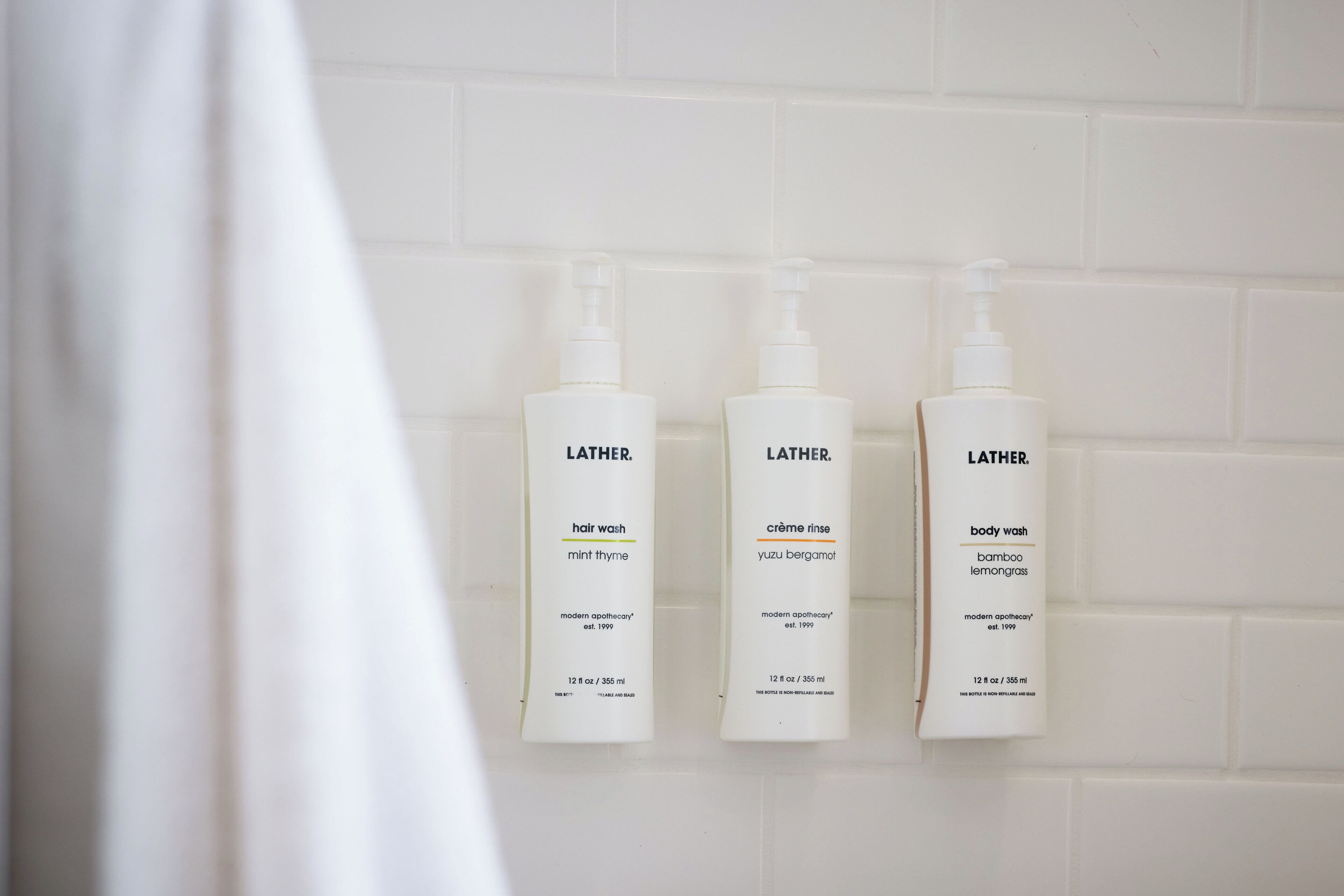 Três embalagens de sabonete líquido Lather sobre uma parede de azulejos brancos.
