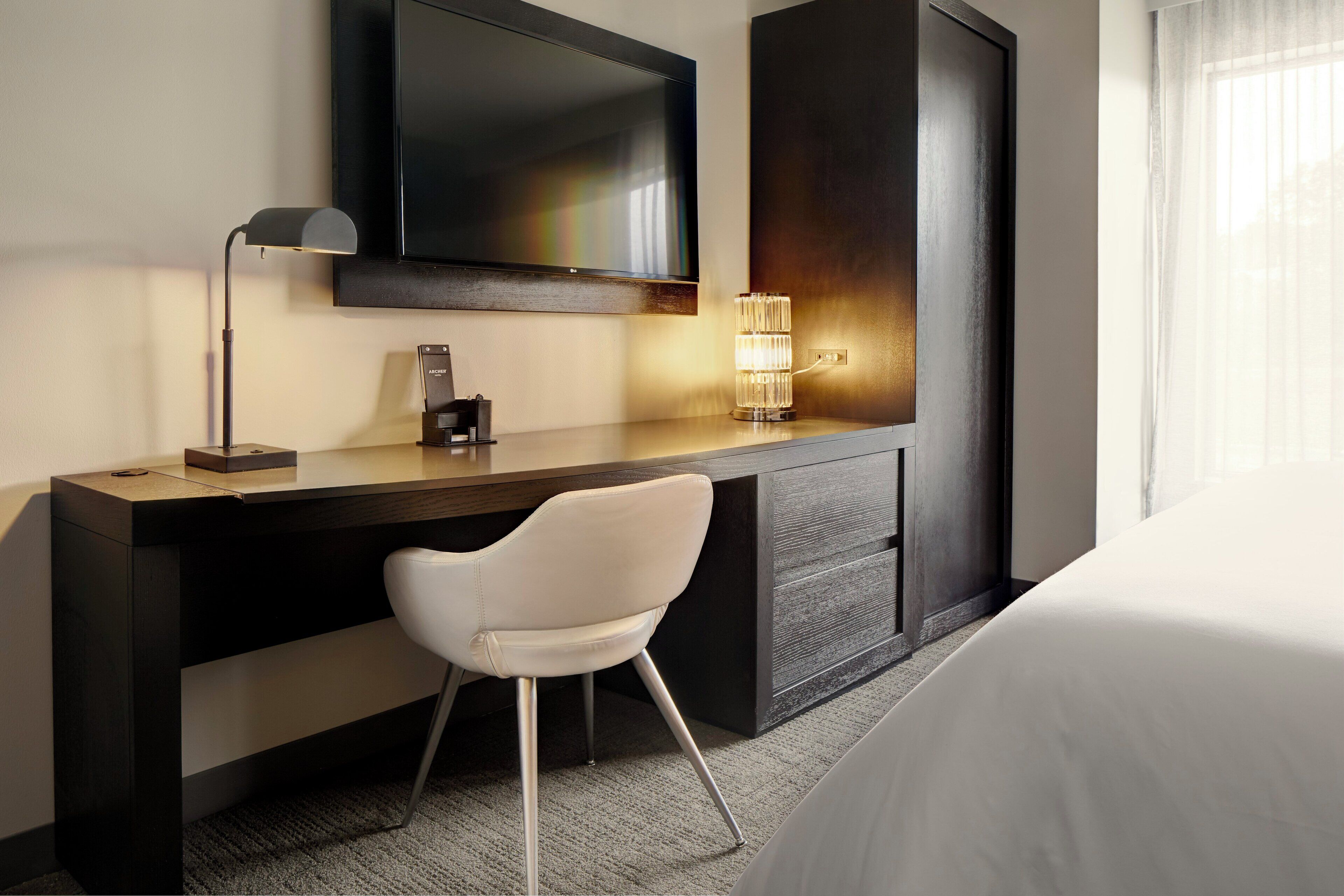 Ein modernes Hotelzimmer mit einem großen Flachbildfernseher an der Wand, einem Schreibtisch mit Lampe, einem weißen Stuhl und einem Bett mit weißer Bettdecke.