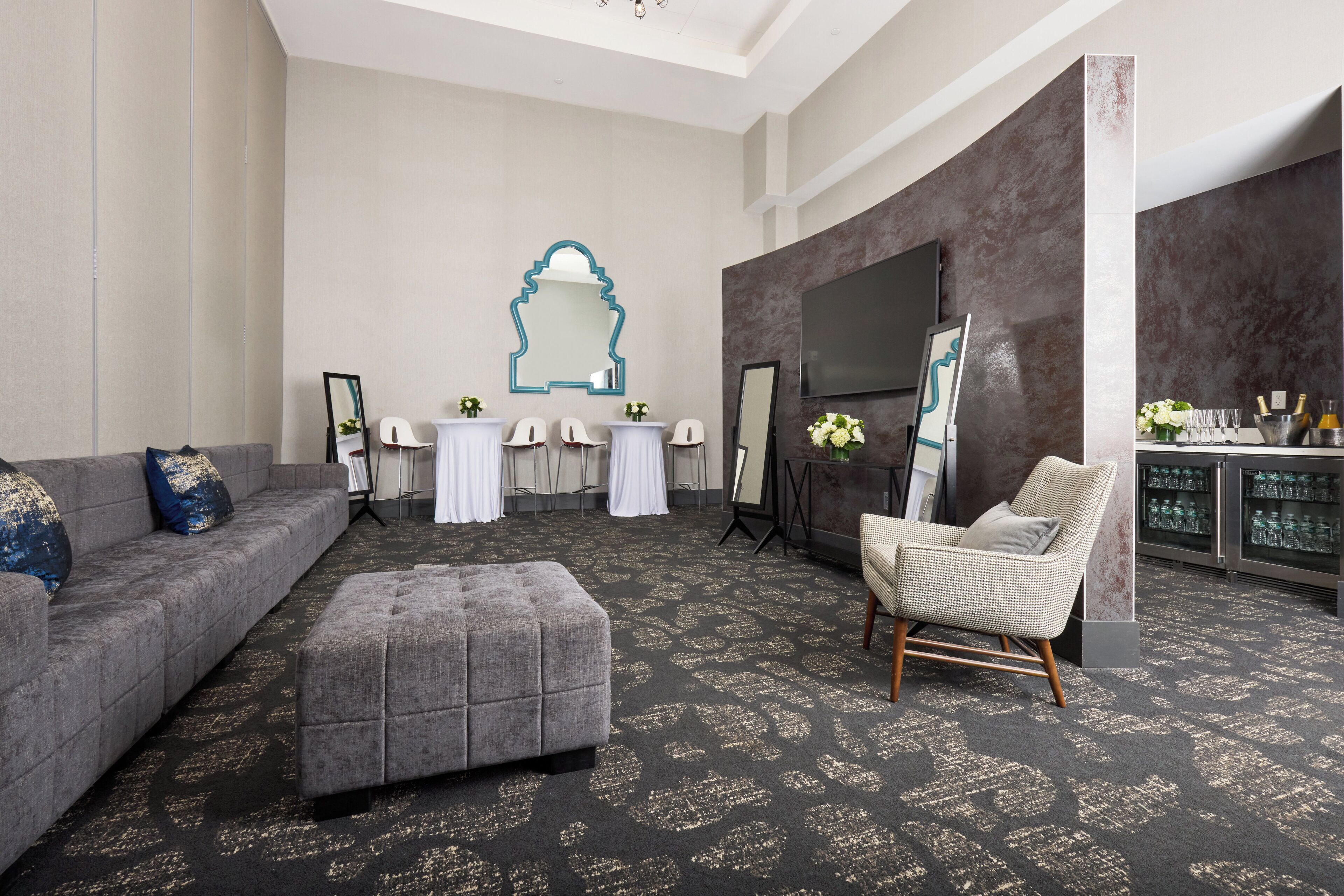A modern hotel room with a grey sofa, an ottoman, a wicker chair, a flat-screen TV, and a bar area with glasses and bottles.
