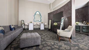 A modern hotel room with a grey sofa, an ottoman, a wicker chair, a flat-screen TV, and a bar area with glasses and bottles.