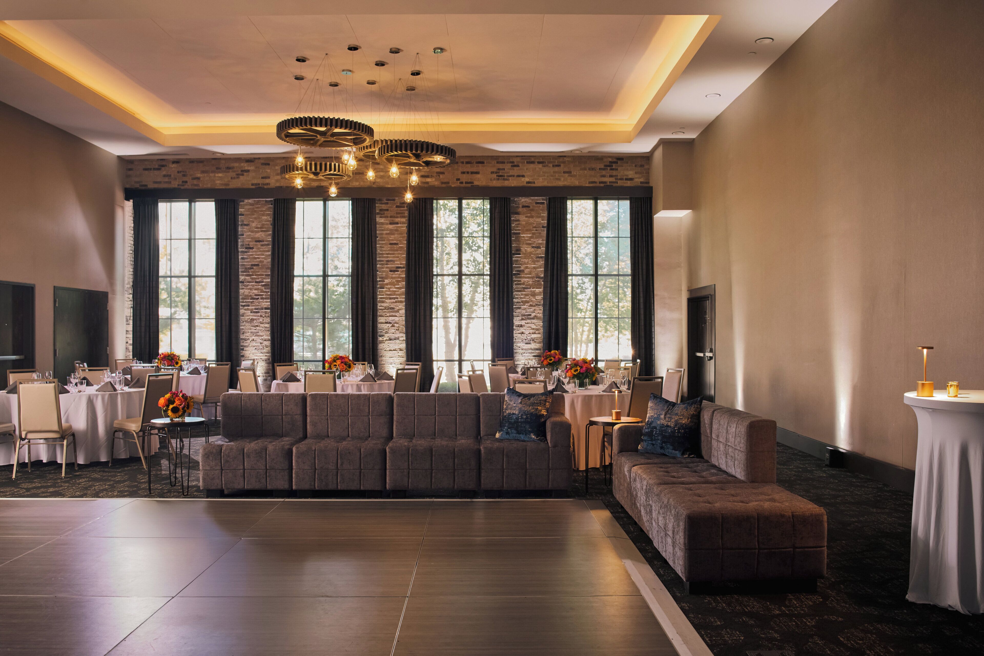 A spacious hotel lobby with a large gray sofa, multiple dining tables set for guests, and a modern chandelier.