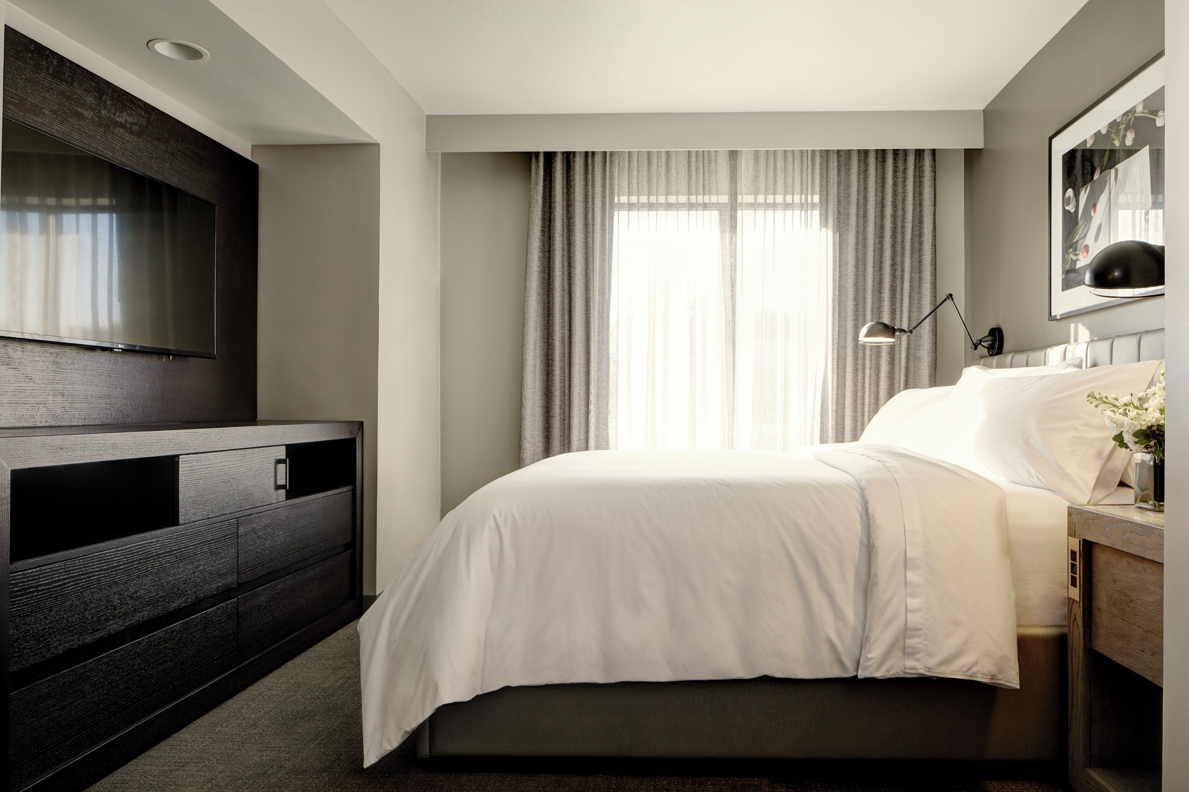 A modern hotel room with a large flat-screen TV mounted on the wall, a bed with white bedding, a bedside table with a lamp, and a window with curtains.