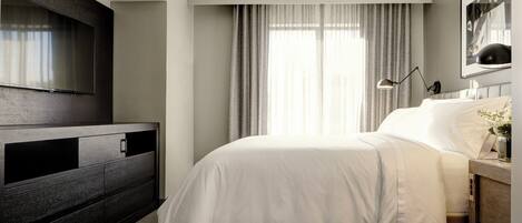 Una habitación de hotel moderna con un televisor de pantalla plana grande montado en la pared, una cama con ropa de cama blanca, una mesita de noche con lámpara y una ventana con cortinas.