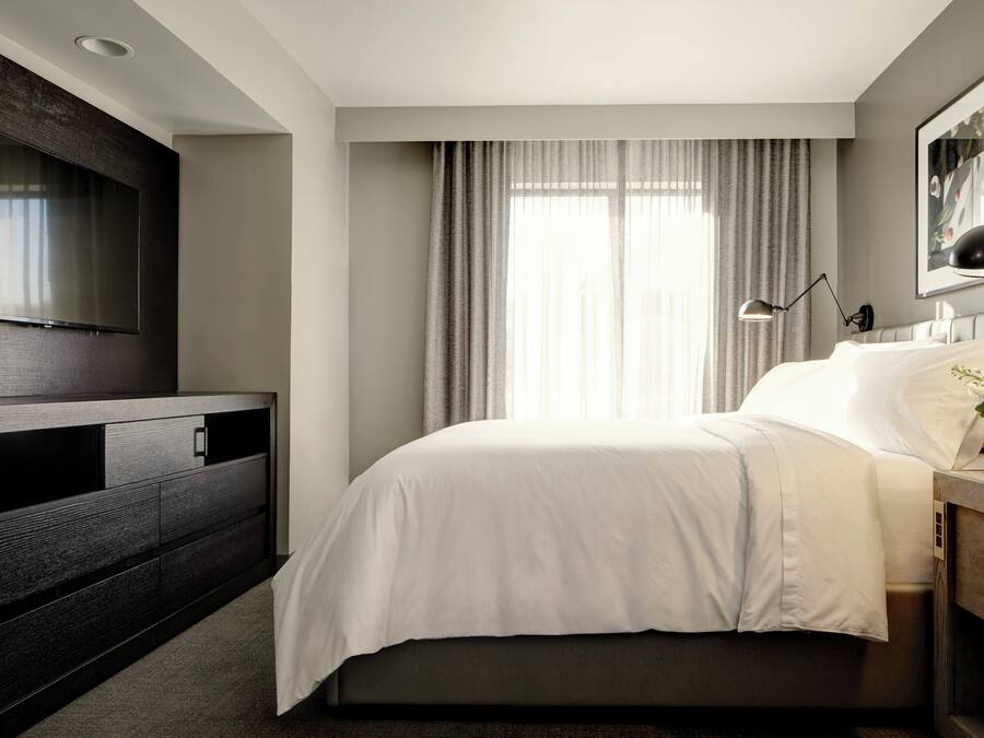 A modern hotel room with a large flat-screen TV mounted on the wall, a bed with white bedding, a bedside table with a lamp, and a window with curtains.