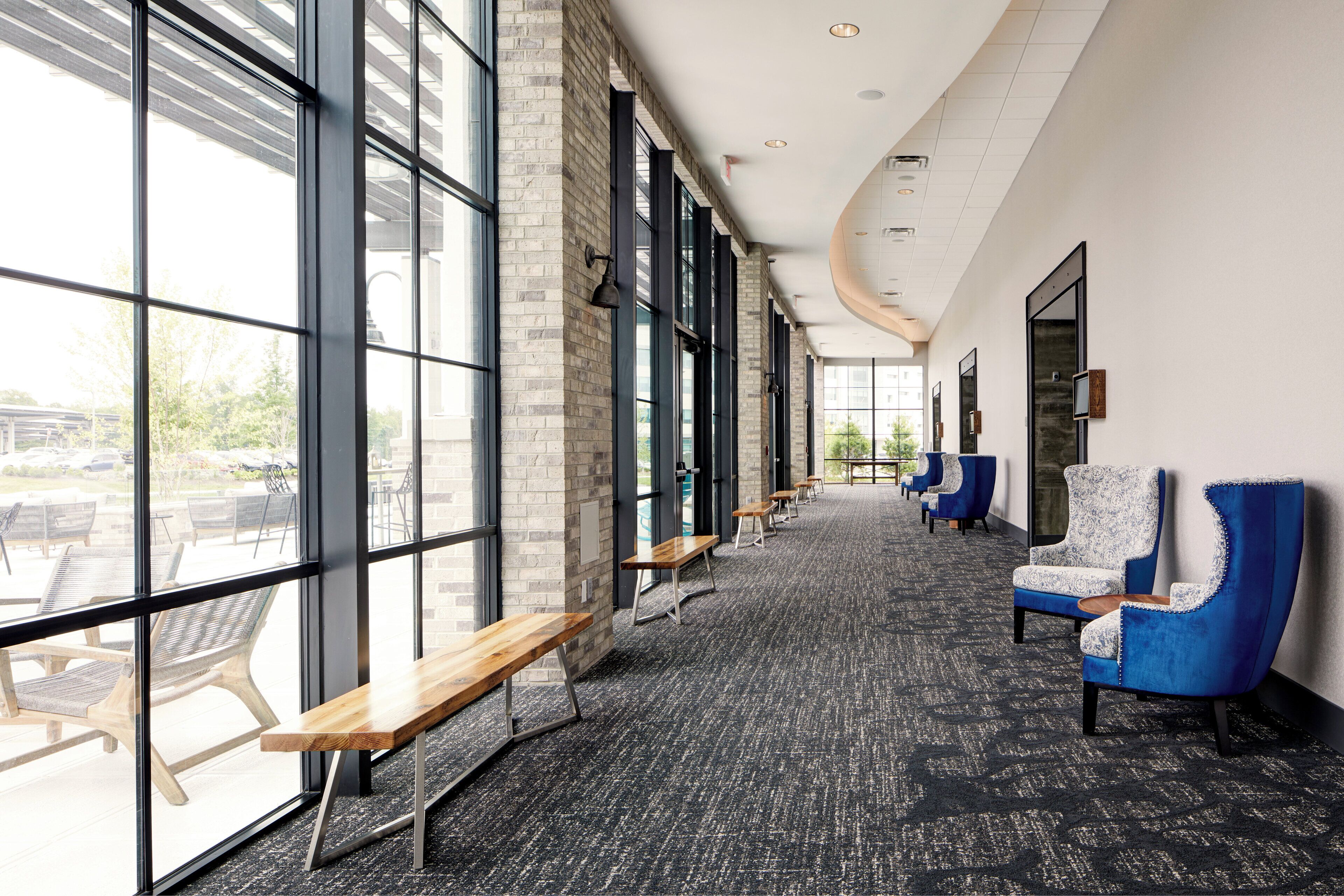 A long corridor with benches and blue chairs, large windows, and a carpeted floor.