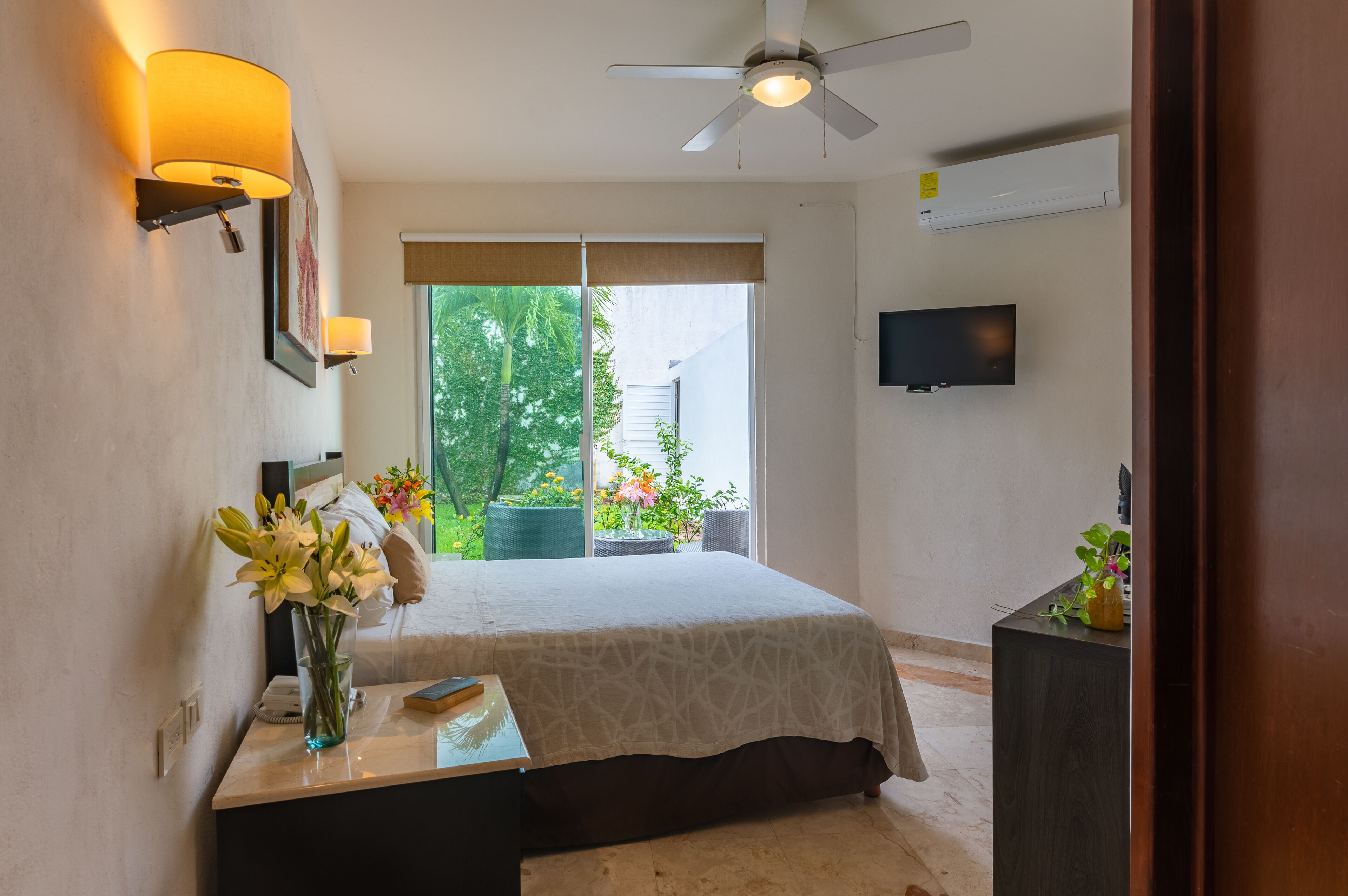 A bedroom with a bed, bedside table, nightstand, TV, ceiling fan, and a view of greenery outside.