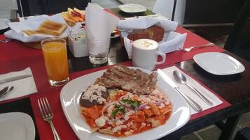A dining table with a plate of pasta, a steak, and a glass of orange juice.