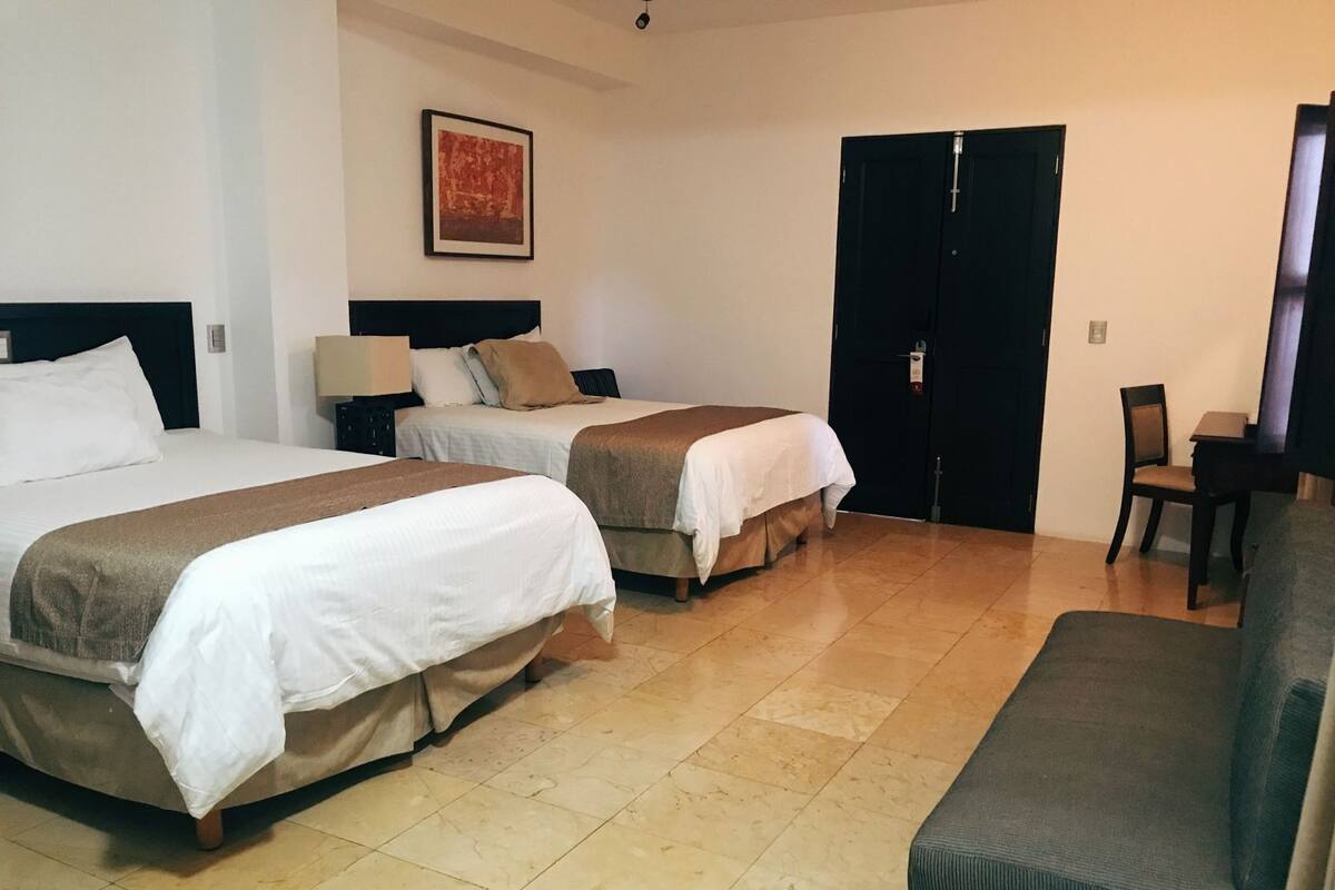 Une chambre d’hôtel avec deux lits, un canapé, une chaise, une porte et un tableau encadré au mur.
