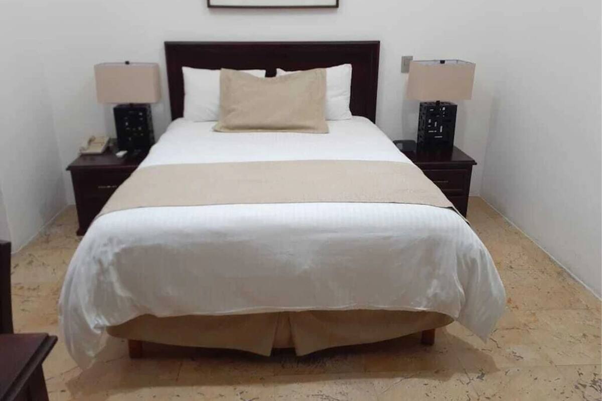 Un lit bien fait, recouvert d’une couette blanche, d’un plaid beige et d’un seul oreiller beige. Deux tables de chevet, chacune surmontée d’une lampe, se trouvent de part et d’autre du lit.