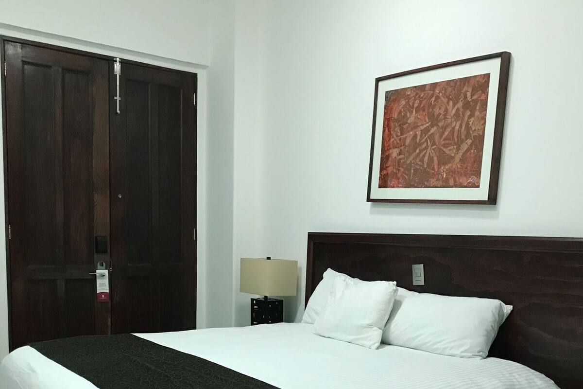 Une chambre d’hôtel avec un lit, une table de chevet, une lampe, une porte et un tableau encadré au mur.