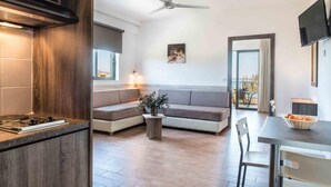 A modern kitchen with a built-in stove, a dining area with a table and chairs, a sofa, and a ceiling fan. - Gerona Mare (Platanias)