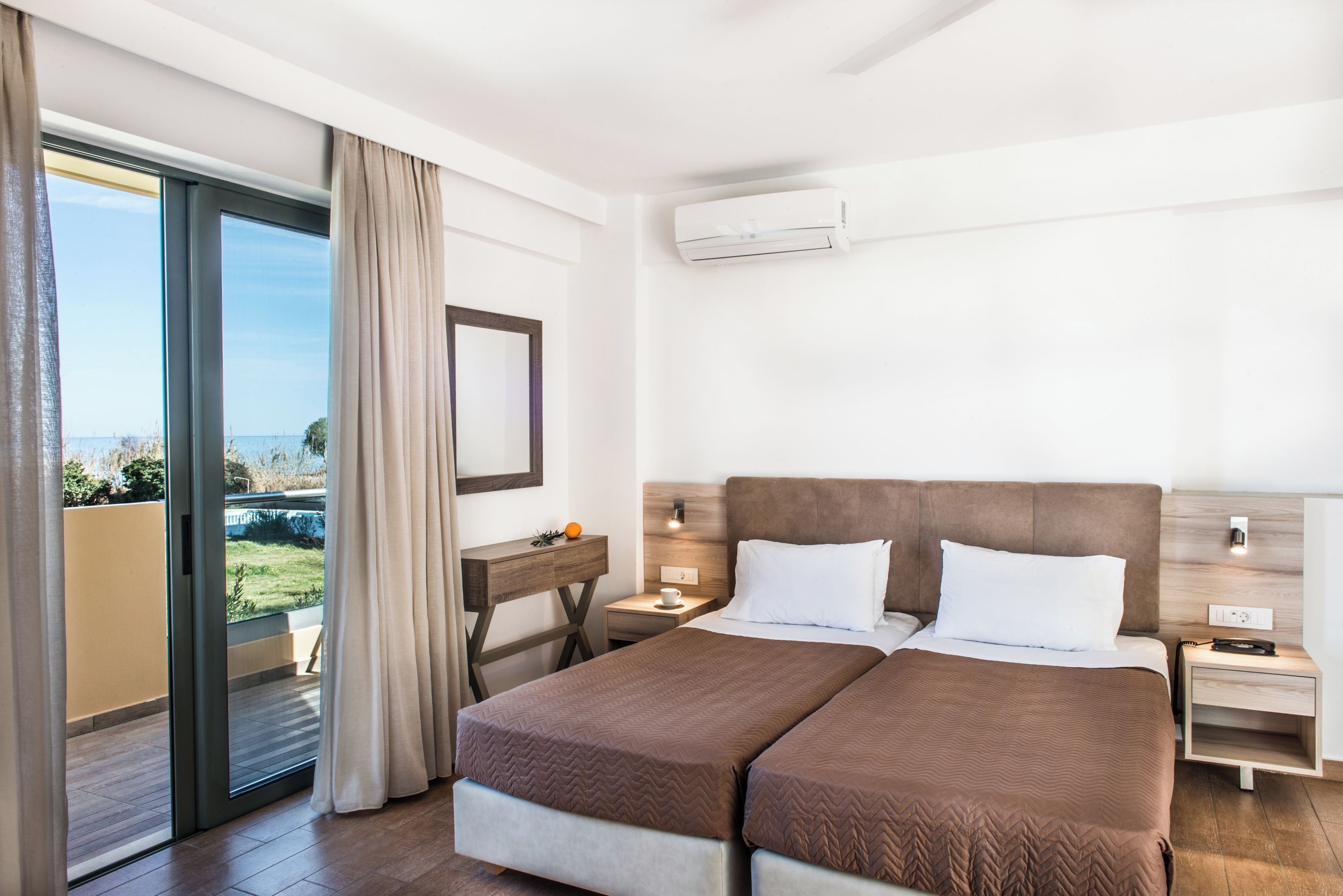A hotel room with a double bed, a bedside table, a nightstand, a mirror, a window with curtains, and an air conditioning unit.