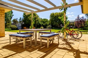 Un patio exterior con mesa y bancos, una bicicleta y un jardín al fondo.