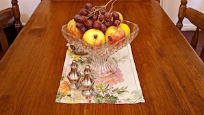Una mesa de comedor de madera con un frutero en el centro, un mantel individual floral y saleros y pimenteros. Detrás de la mesa hay un aparador de madera con platos azules y blancos y un letrero de Arnott's Biscuits.