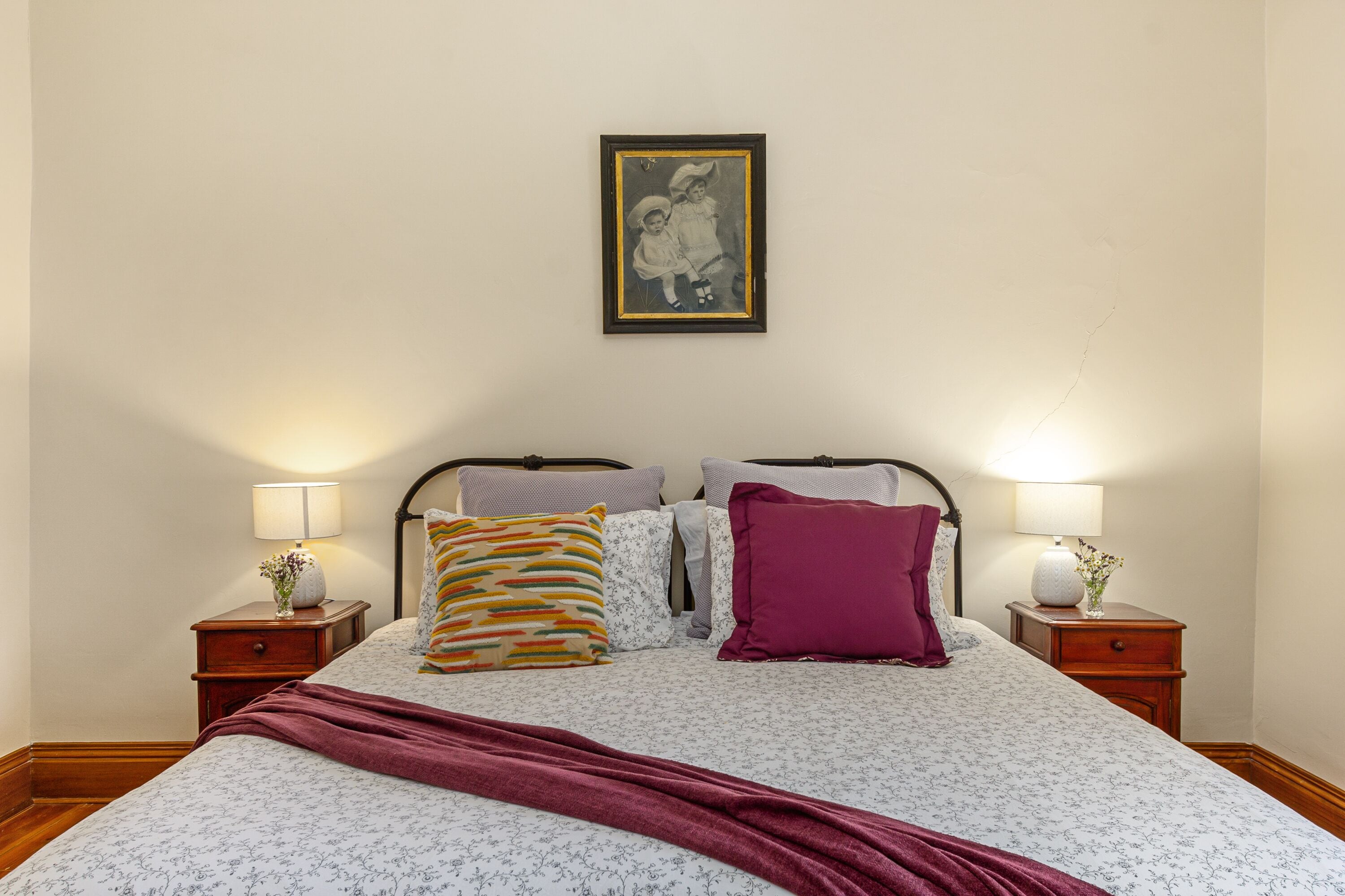 A neatly made bed with a floral bedspread, two pillows, a red throw blanket, and a framed picture above it. There are two bedside tables with lamps and decorative vases.