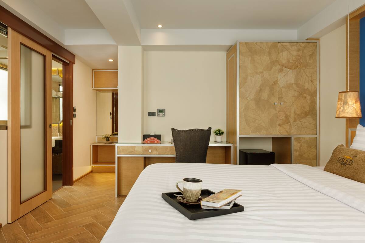 A hotel room with a large bed, a desk, and a chair. There is a tray with a cup and a book on the bed.