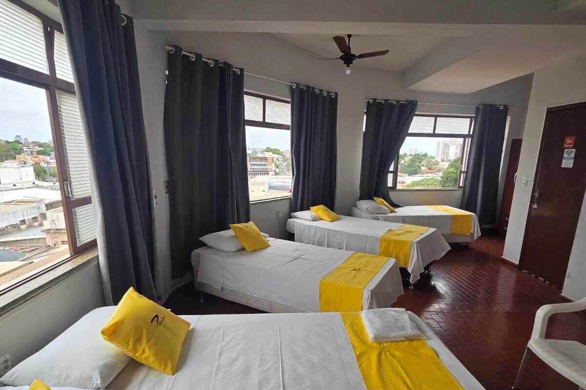 A hotel room with three beds, each with white bedding and yellow accents. There are large windows with curtains, a ceiling fan, and a view of the cityscape.