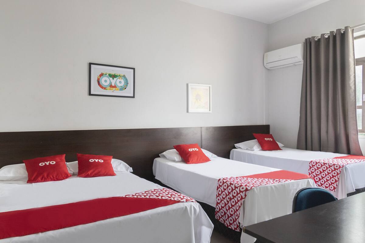 A hotel room with two beds, each with a red OYO branded pillow, white bedding, and a red and white patterned bedspread. There is a framed picture on the wall, an air conditioning unit, and a window with curtains.