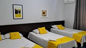 Habitación de hotel con tres camas individuales, cada una con colcha y almohada amarillas, ropa de cama blanca y un ventilador de techo. Hay dos cuadros en la pared, una ventana con cortinas y un equipo de aire acondicionado.