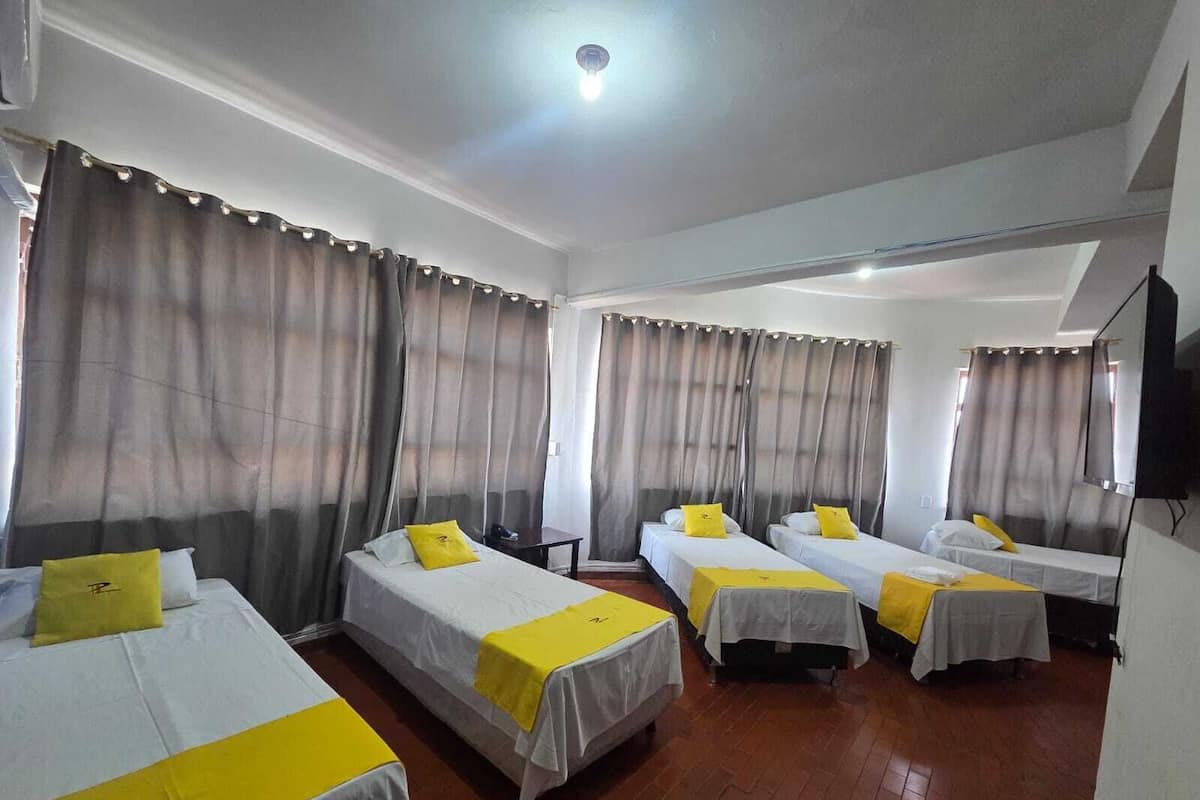 A hotel room with four single beds, each with a yellow bedspread and pillow. The room has a wooden floor, white walls, and large curtains.