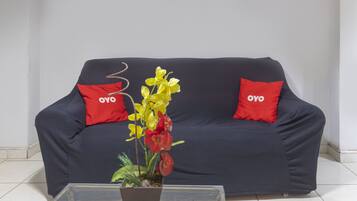 Um sofá azul escuro com duas almofadas vermelhas, cada uma com o logotipo "OYO". À frente do sofá, uma mesa de centro de vidro transparente com uma planta em vaso.