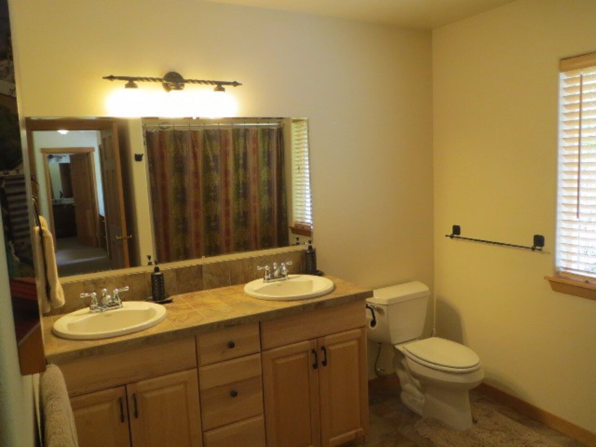 A bathroom with two sinks, a toilet, a mirror, and a window with blinds.